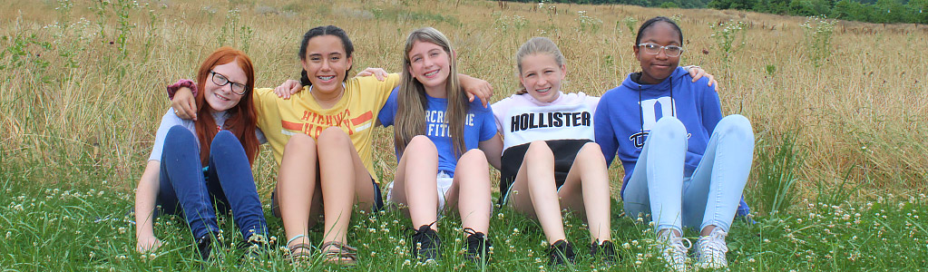 5 middle school girls sitting in tall grass on a mountain side, with arms wrapped around each other.