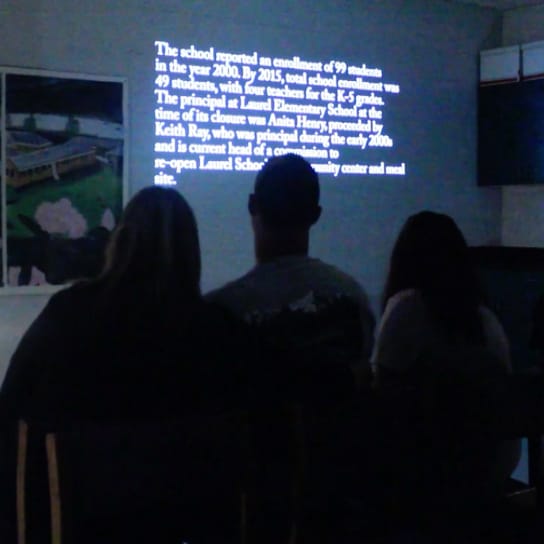A number of people in a dark room watching a projected image on a school room wall. The image is from a PAGE installation.