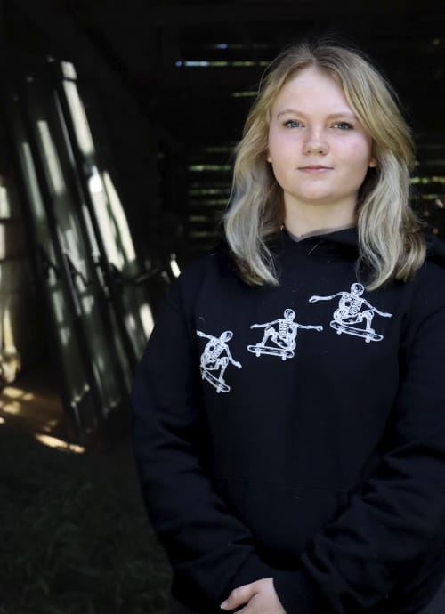 Katlyn, a young teen, looks confidently into the camera with her hands clasped in front of her. She's wearing a black sweatshirt and standing in the doorway of an old barn.