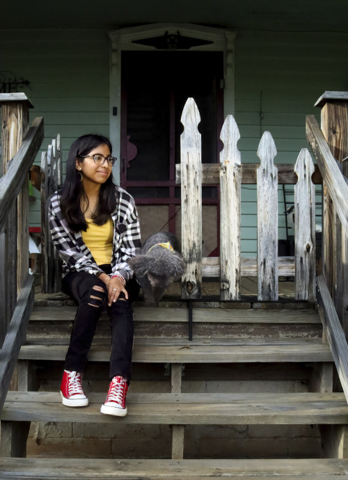 Maria, a young teen, sits on weathered front porch steps with a small dog. She's wearing bright red converse sneakers and a flannel shirt. She's looking off into the distance with a slight smile.