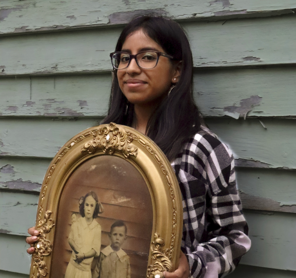 Maria stands against the side of her house, weathered siding behind her. She's holding an antique photo of two young children in an ornate gold frame.